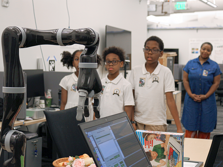 three small students looking at a robotic arm work, with their teacher in the background
